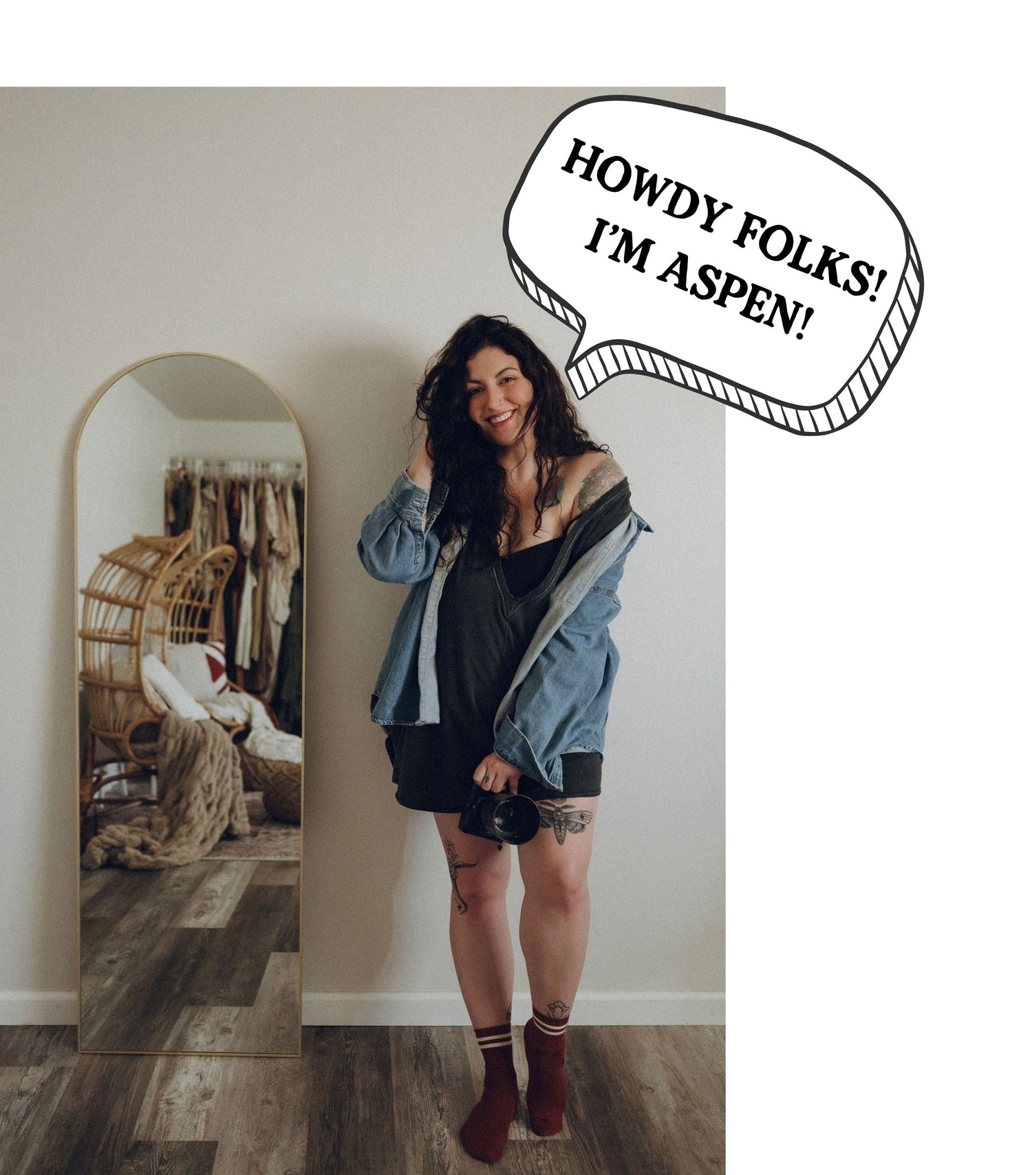 A woman stands indoors wearing a black dress, an open light blue denim jacket, and dark socks, with a large speech bubble reading 'HOWDY FOLKS! I'M ASPEN!' overlaid on the top right. A tall arched mirror on the left reflects part of a room with a hanging chair and clothing.